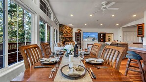 Un coin repas avec une table en bois dressée pour six personnes, entourée de chaises, avec une cheminée en pierre et une grande fenêtre.