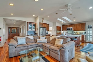A spacious living area with a sectional sofa, a glass coffee table, wooden flooring, and a kitchen with wooden cabinets and modern appliances.
