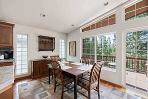 A dining area with a wooden table and chairs, a kitchen with wooden cabinets, and large windows offering a view of trees.