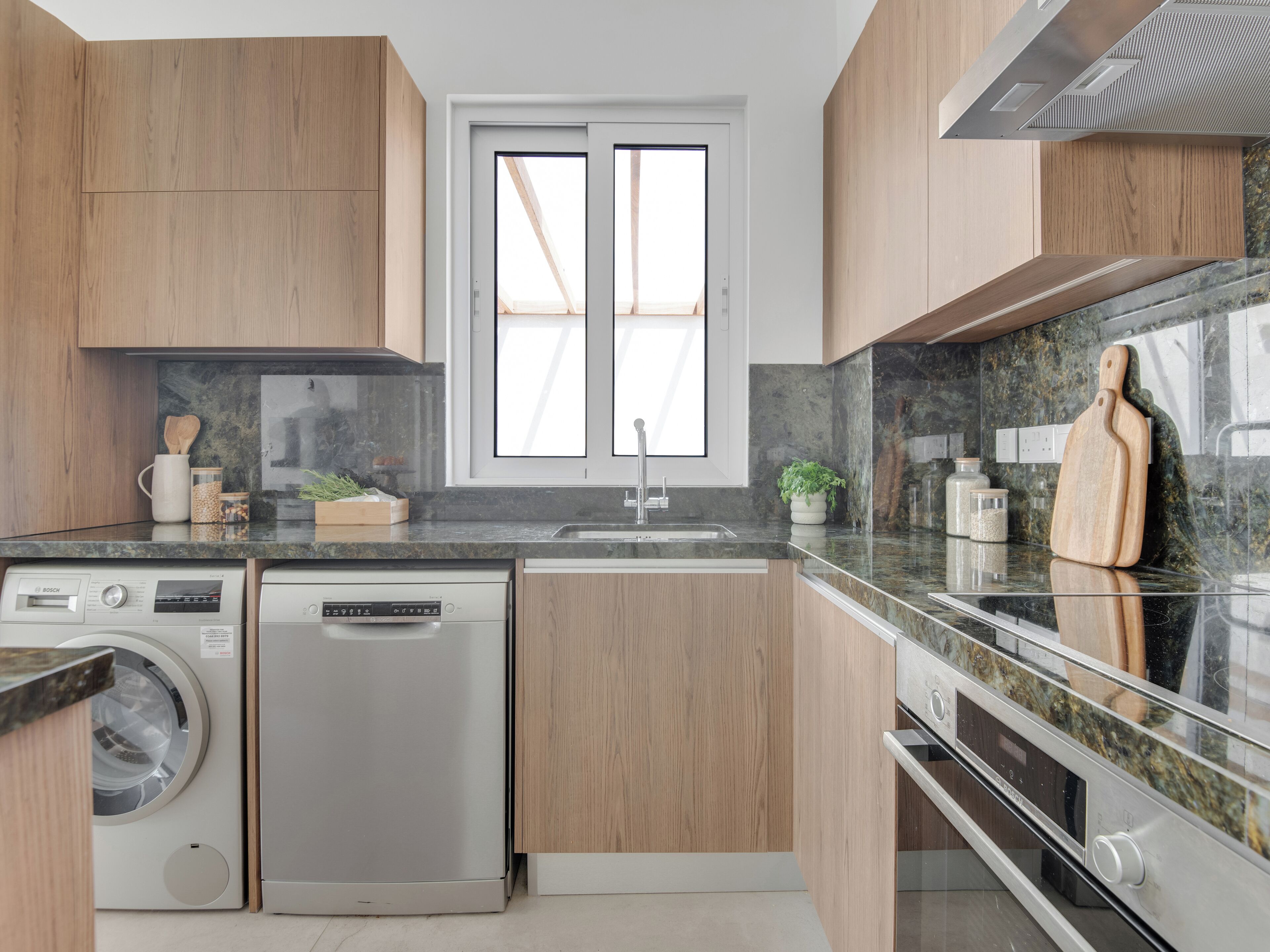 A modern kitchen with wooden cabinets, a built-in dishwasher, a sink, and a window with blinds.