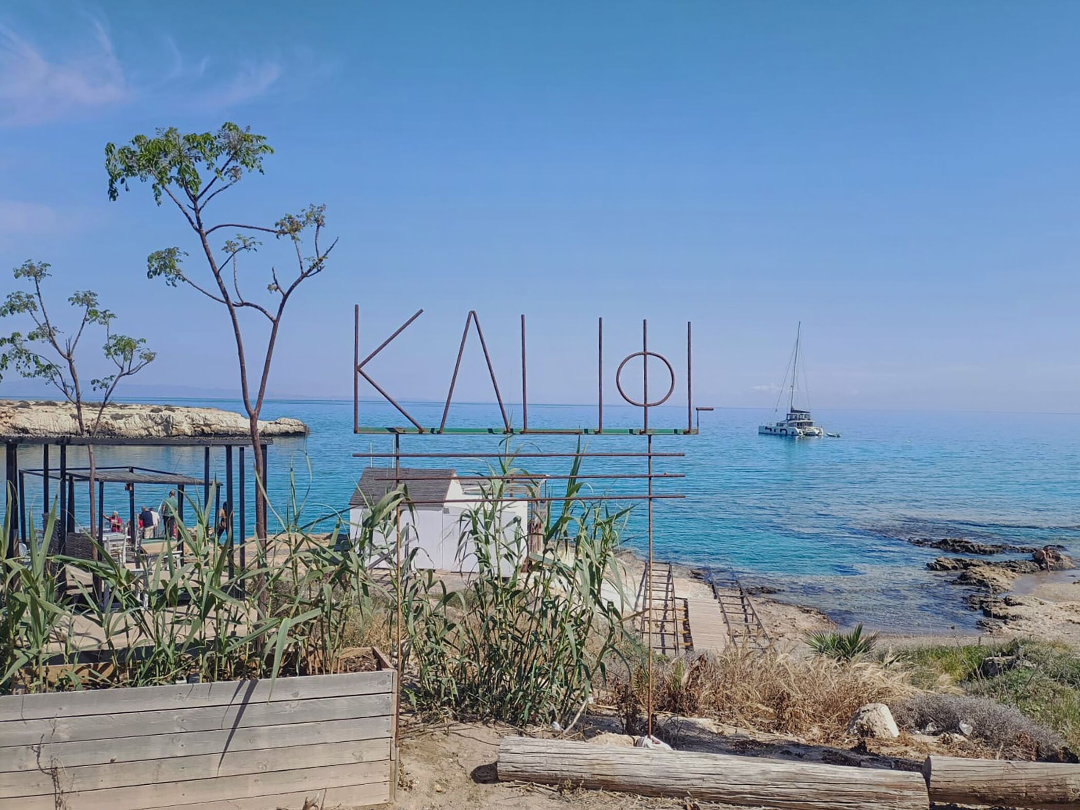 Une plage avec une structure en bois, un voilier et une eau d’un bleu limpide.