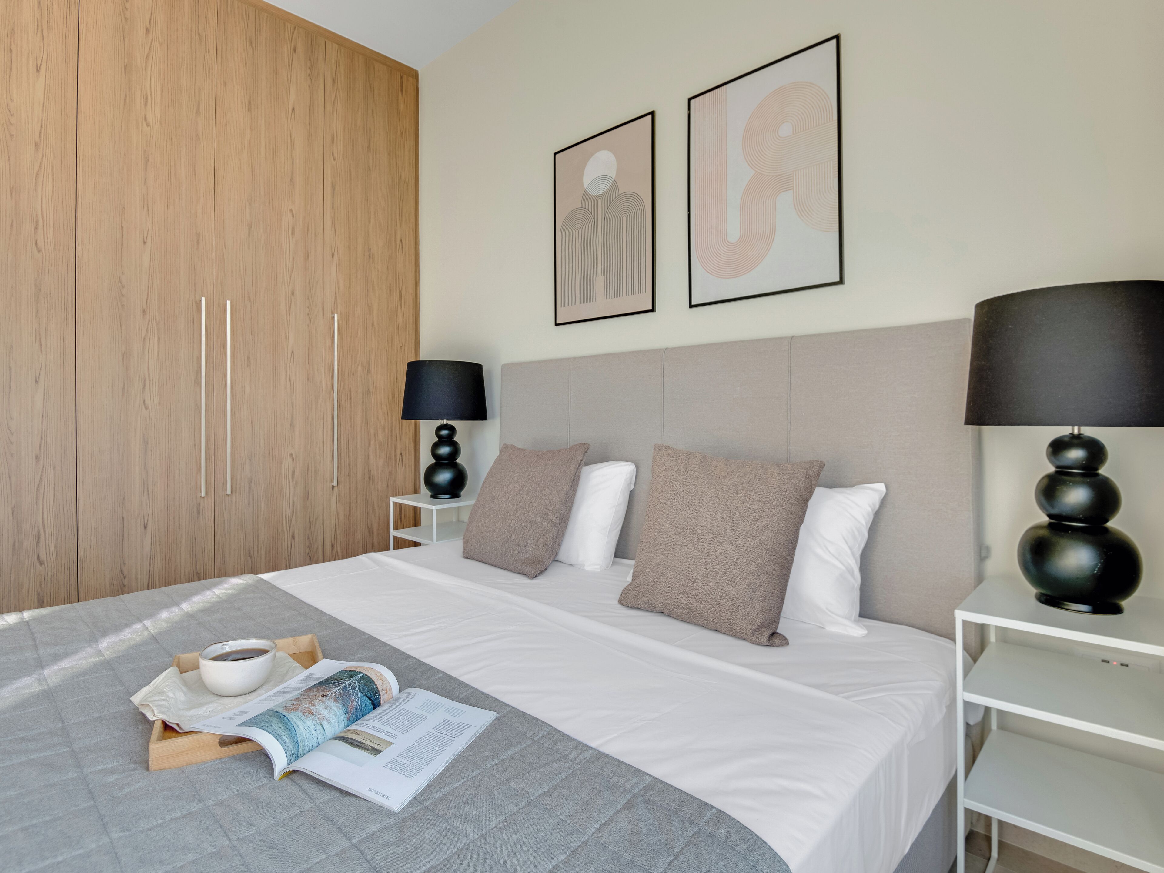 A modern hotel room with a bed, a bedside table with a lamp, a wardrobe, and artwork on the wall.