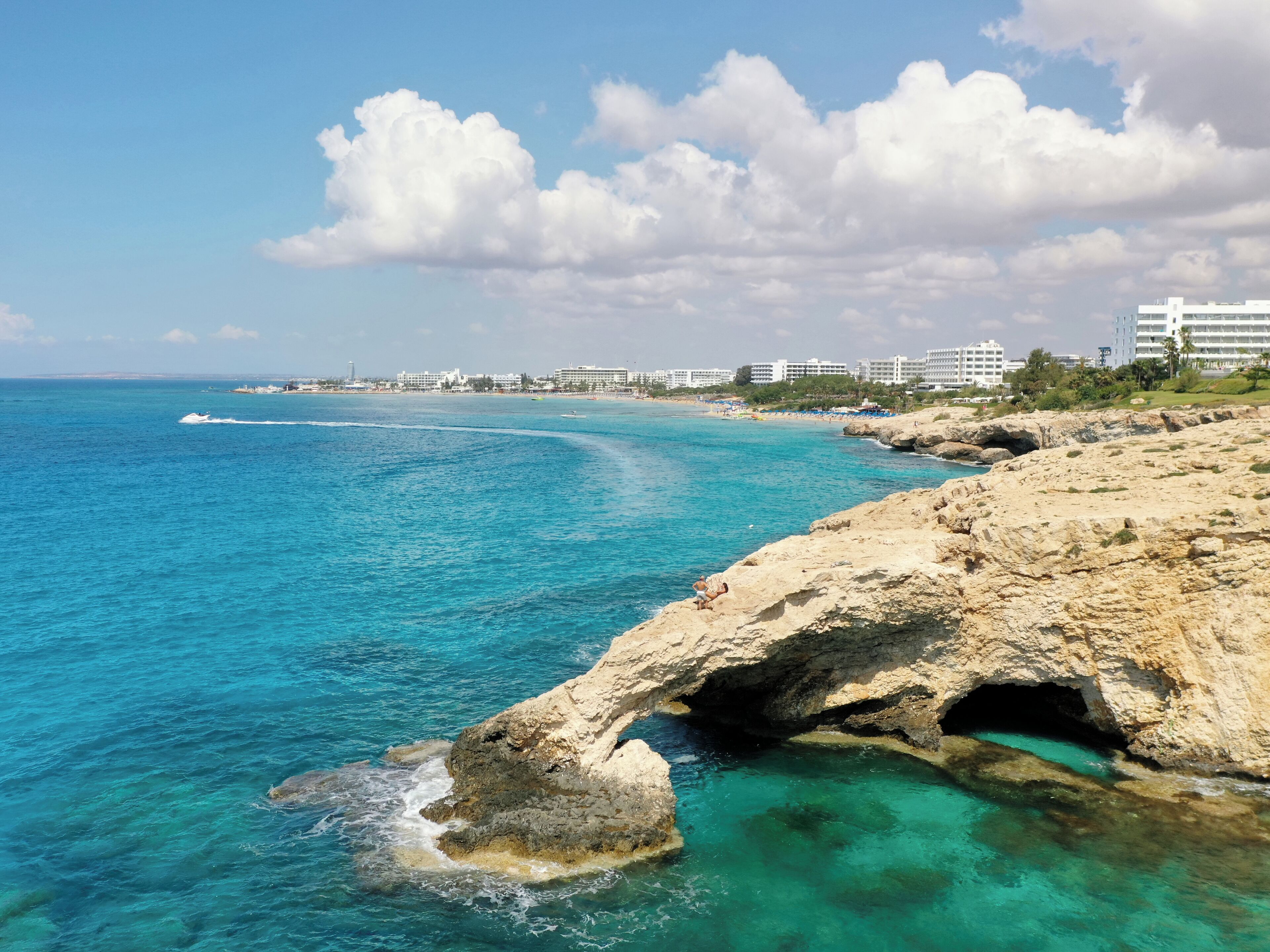 A coastal scene with clear turquoise waters, a rocky shoreline, and a distant cityscape.