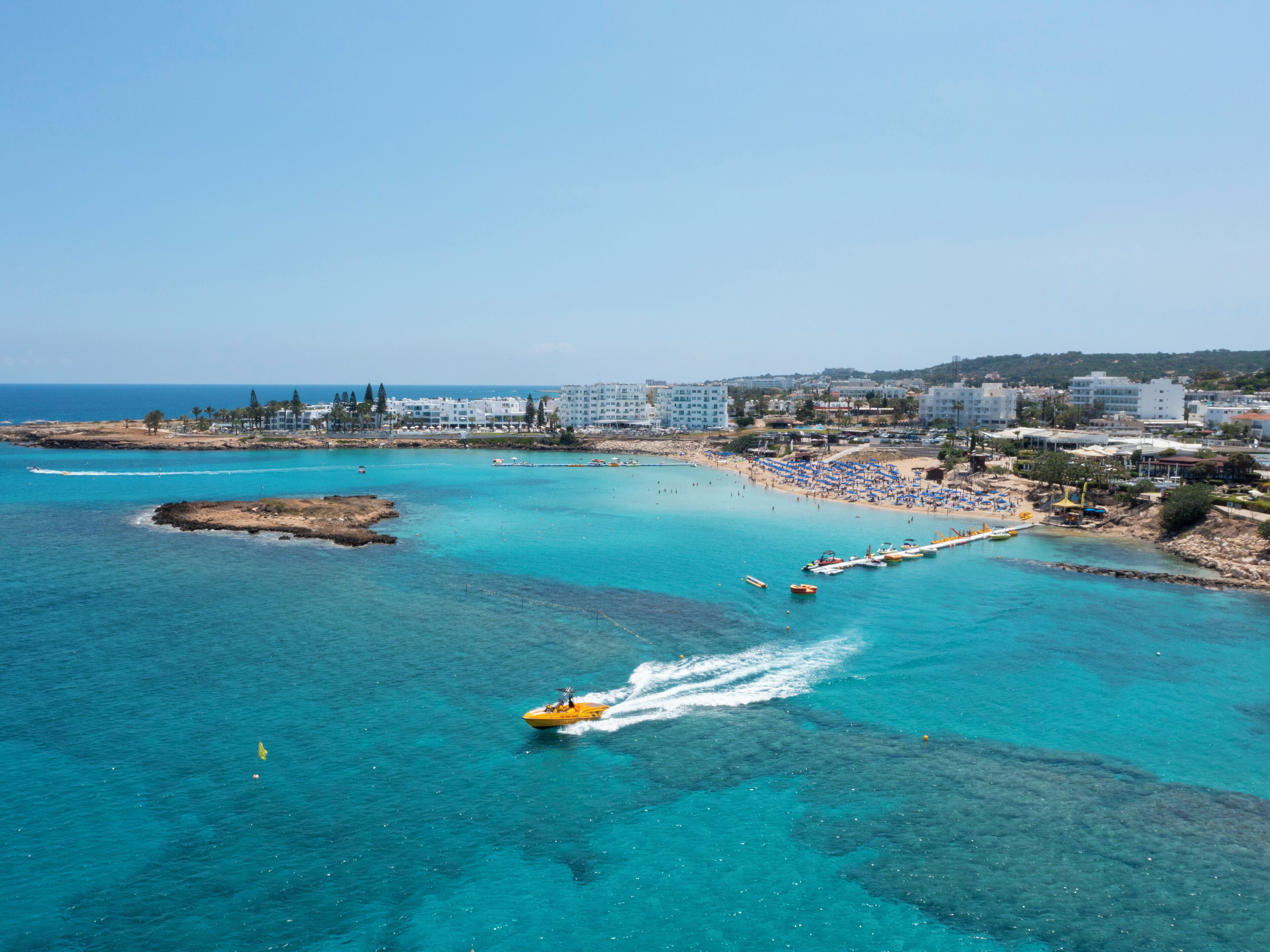 Una zona costiera con mare cristallino, un jet ski e una spiaggia con ombrelloni e persone.
