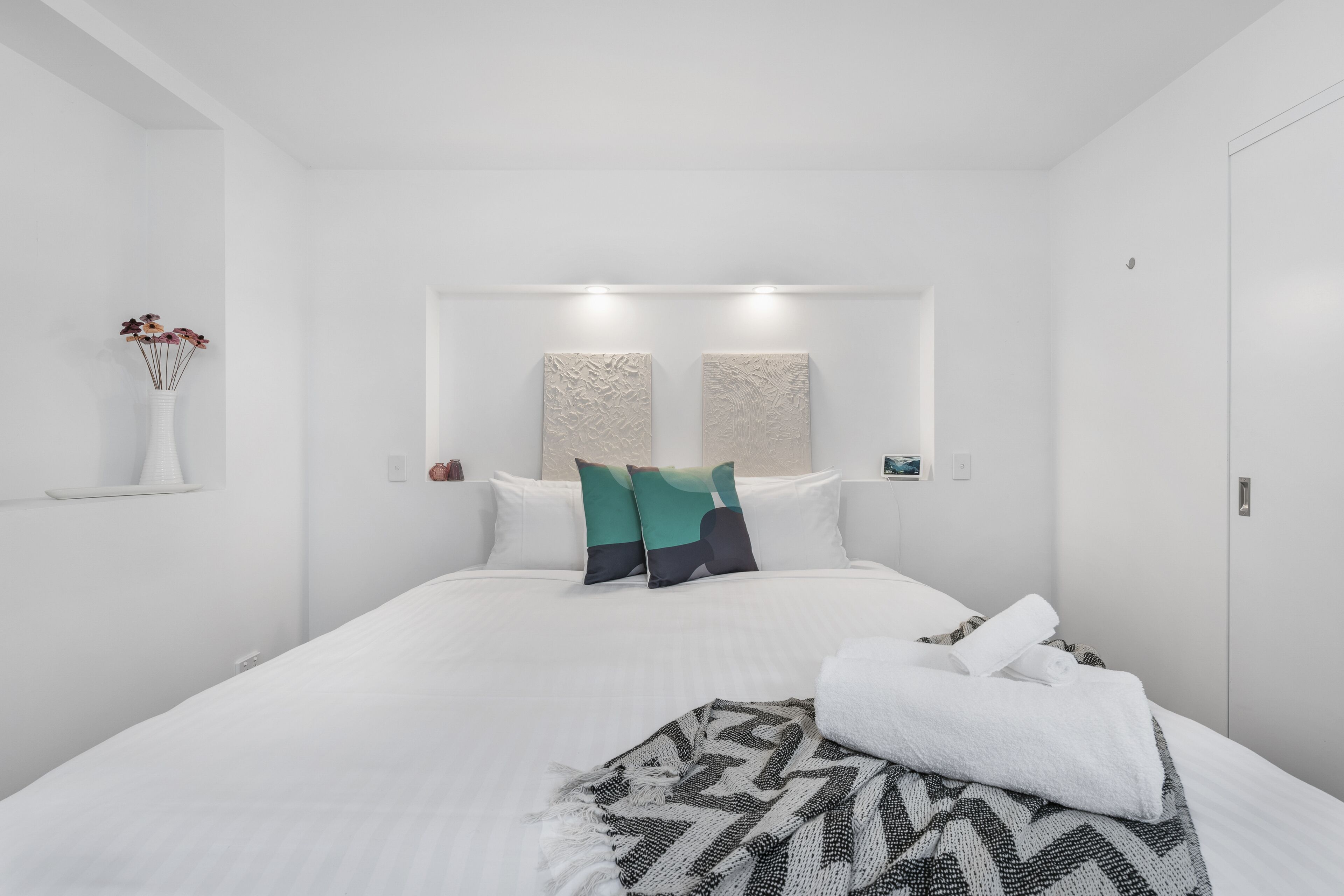 Een moderne hotelkamer in wit kleurenschema, met een opgemaakt bed, een decoratief wandkunstwerk en een vaas met bloemen.