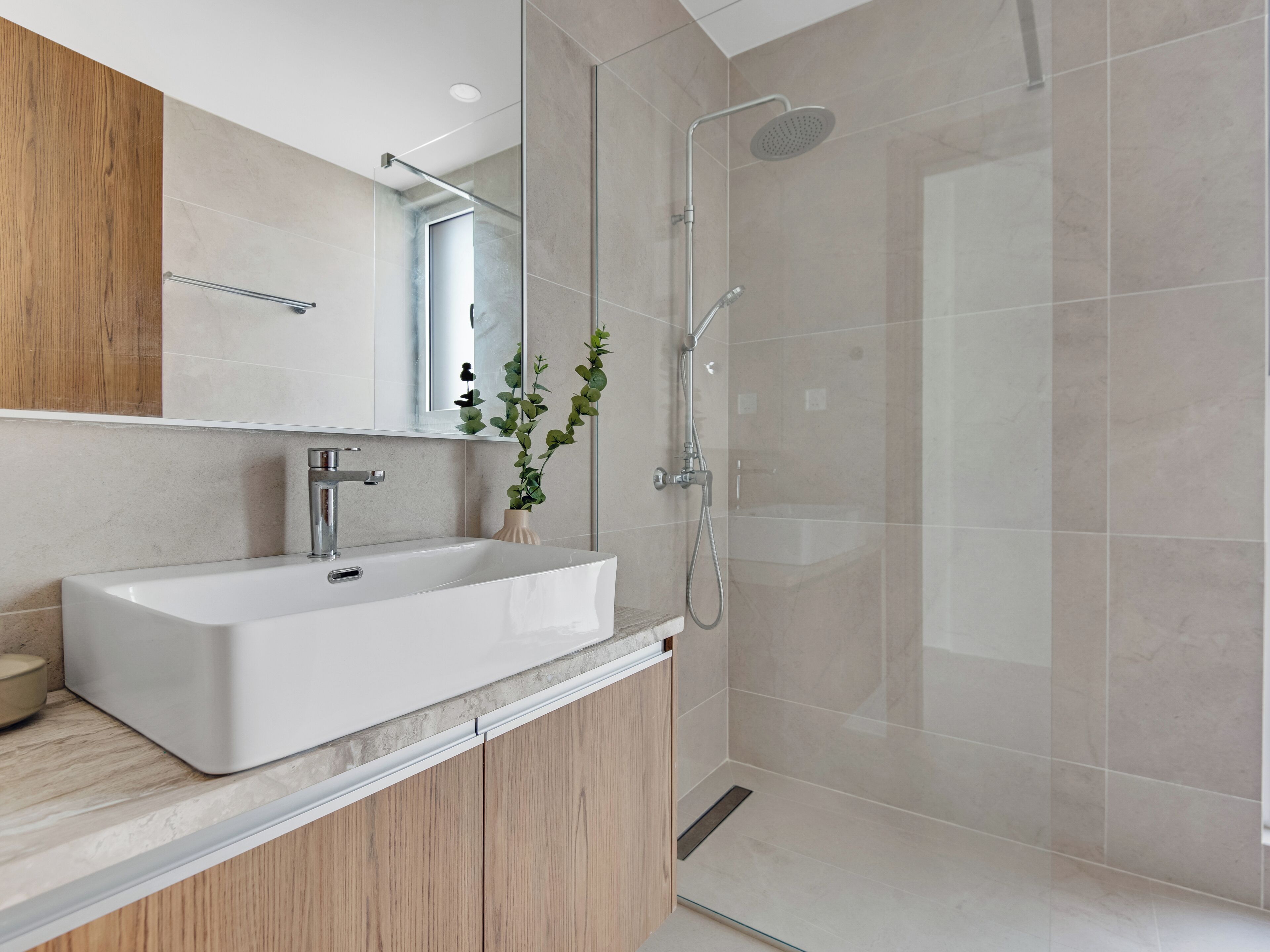 A modern bathroom with a white sink, wooden cabinets, a glass shower enclosure, and a potted plant.