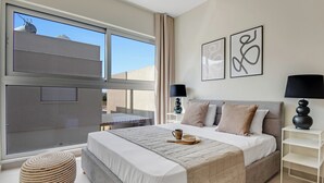 Een moderne slaapkamer met een groot bed, nachtkastjes, een rond pouf en uitzicht op een gebouw door het raam.