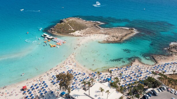 Luchtbeeld van een strand met helder turquoise water, een rotsachtige uitloper en vele parasols.