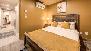 Camera da letto con testiera in legno, letto con biancheria bianca, copriletto giallo, comodino con lampada e condizionatore a muro.