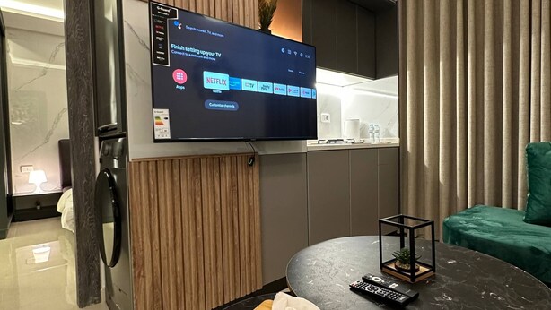 A modern living room with a flat-screen TV mounted on the wall, a round marble table with a remote and tissues, a green sofa, and a washing machine in the background. - JRE Modern Apartment In Abdoun-amman (Amman)
