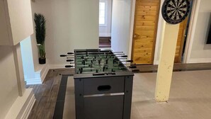 Une table de baby-foot dans une pièce au plancher de bois, avec un jeu de fléchettes et une porte en bois.