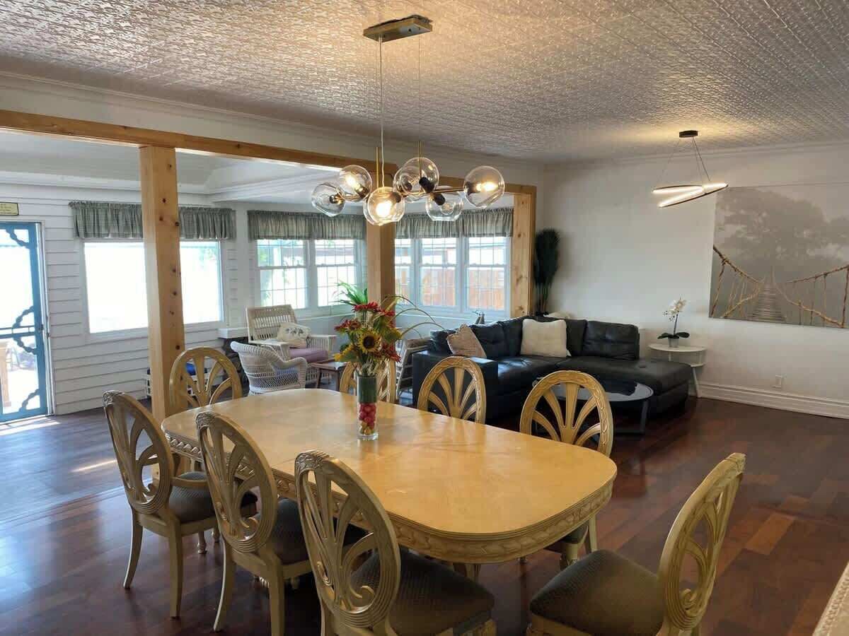 A spacious dining area with a wooden table and chairs, a chandelier, and a view of the living room with a sofa and a painting on the wall.