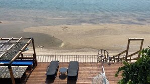Une terrasse en bois avec des chaises longues et une table, donnant sur une plage de sable fin et une mer calme.