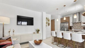 Eine moderne Küche mit weißen Schränken, einer zentralen Kochinsel mit Barhocker und einem an der Wand montierten Flachbildfernseher.