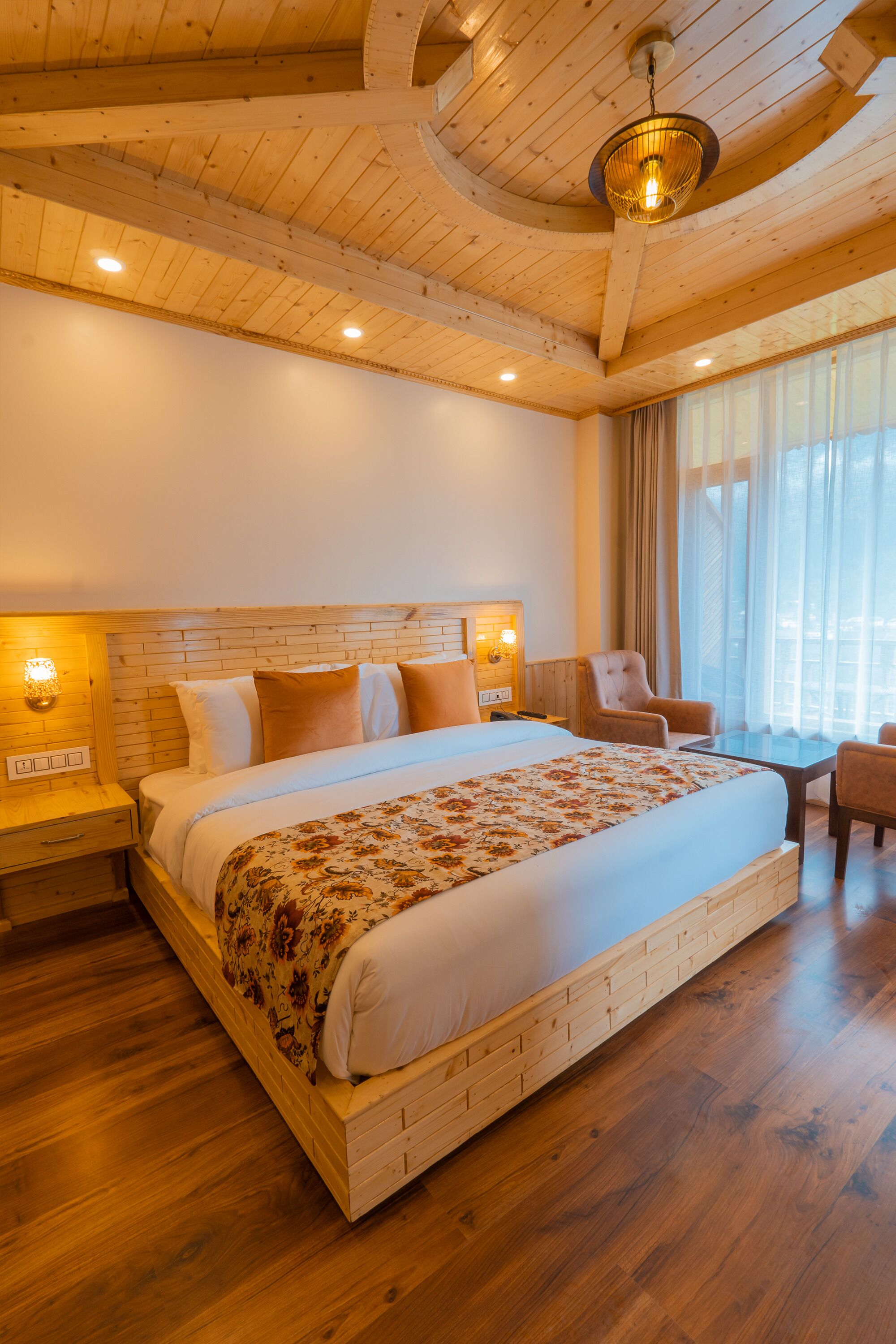 Un lit en bois avec une couvre-lit à motifs floraux, une table de chevet avec une lampe et une chaise près d’une fenêtre ornée de rideaux.