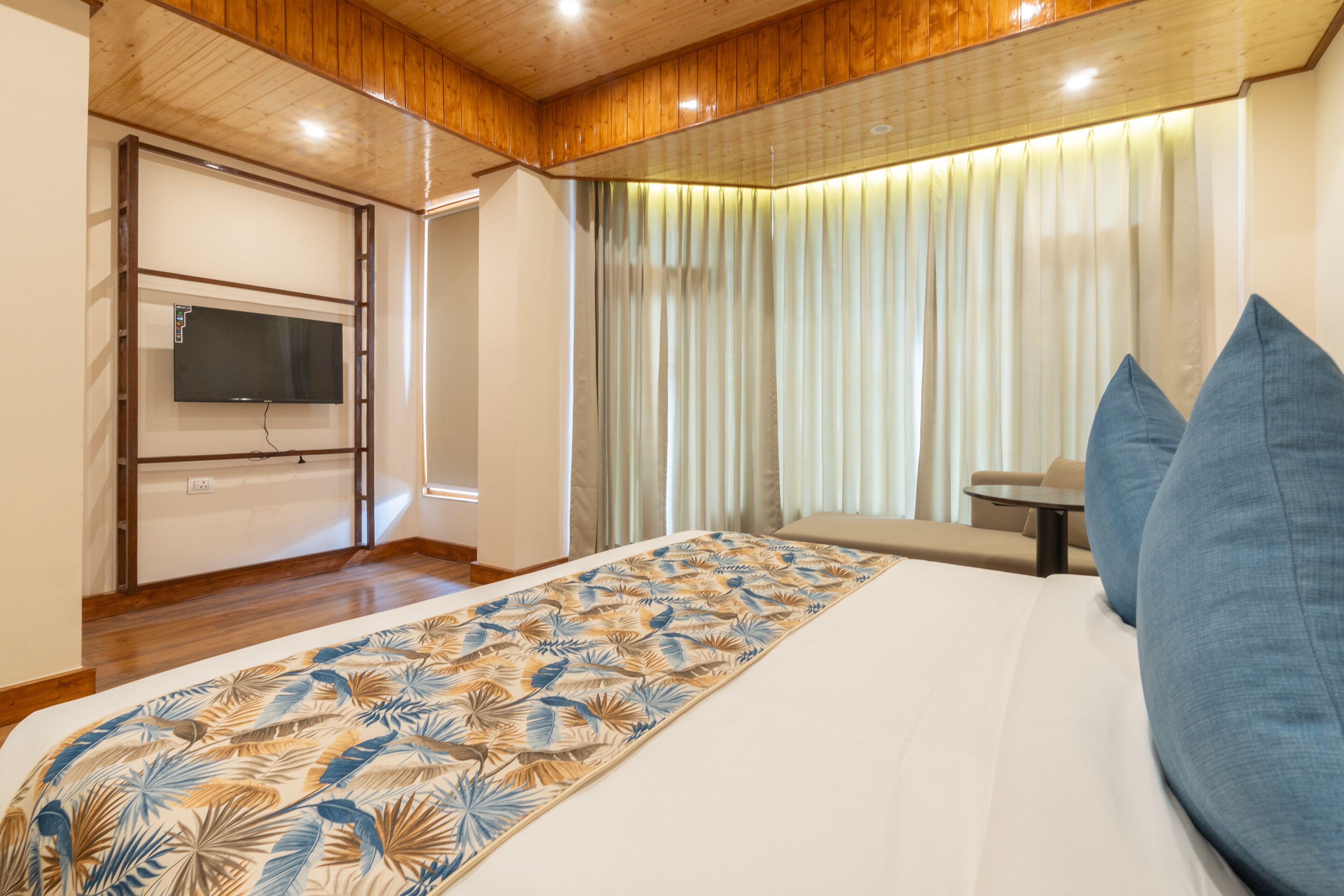 A hotel room with a bed, a television mounted on the wall, a window with curtains, and a small table with a chair.