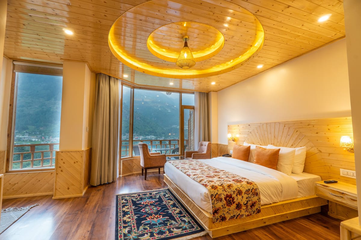 Een ruime slaapkamer met een groot bed, houten plafond en uitzicht op een bergachtig landschap door de ramen.
