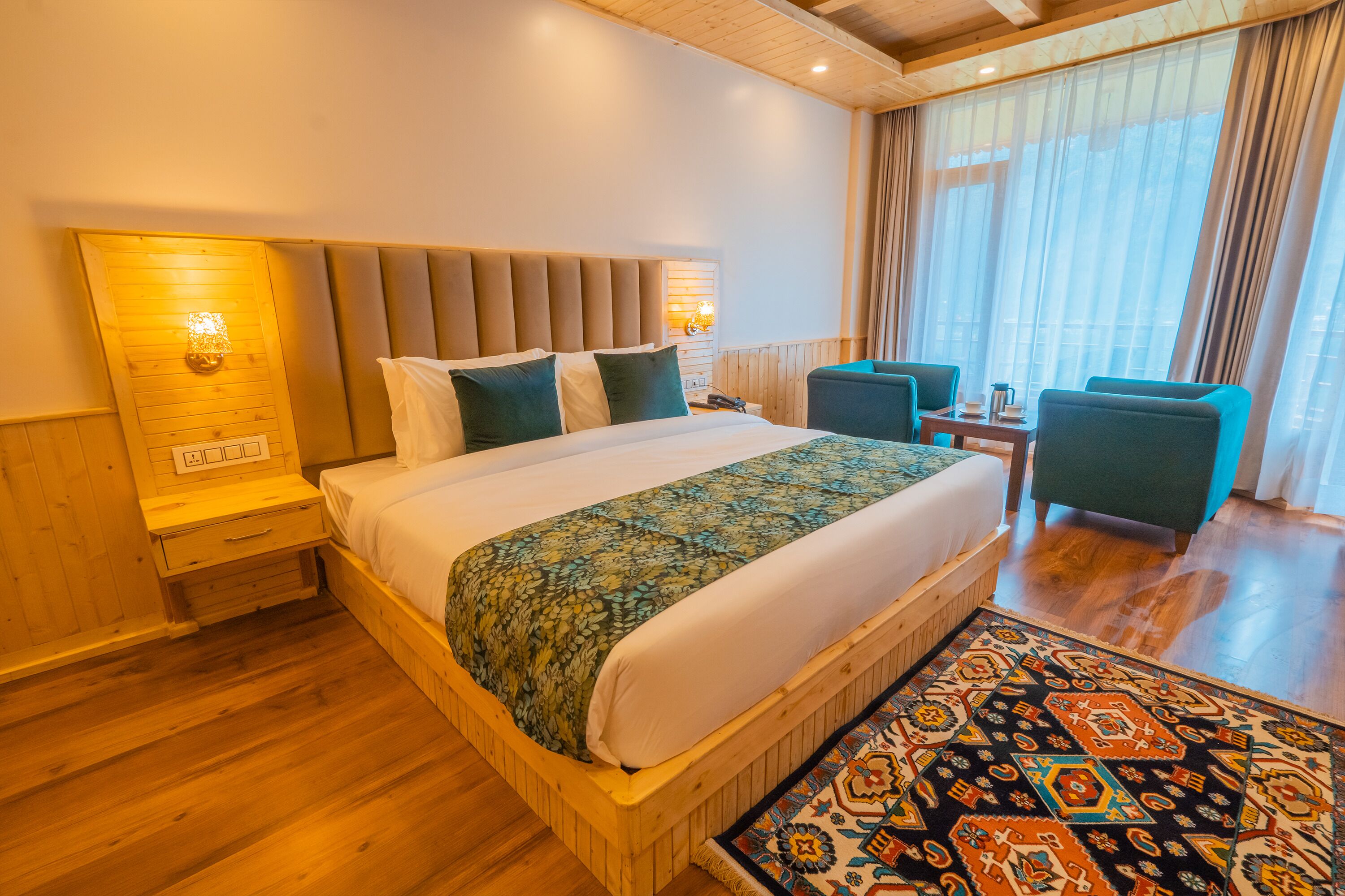 A hotel room with a large bed, a wooden headboard, a nightstand, a desk with two chairs, and a patterned rug.