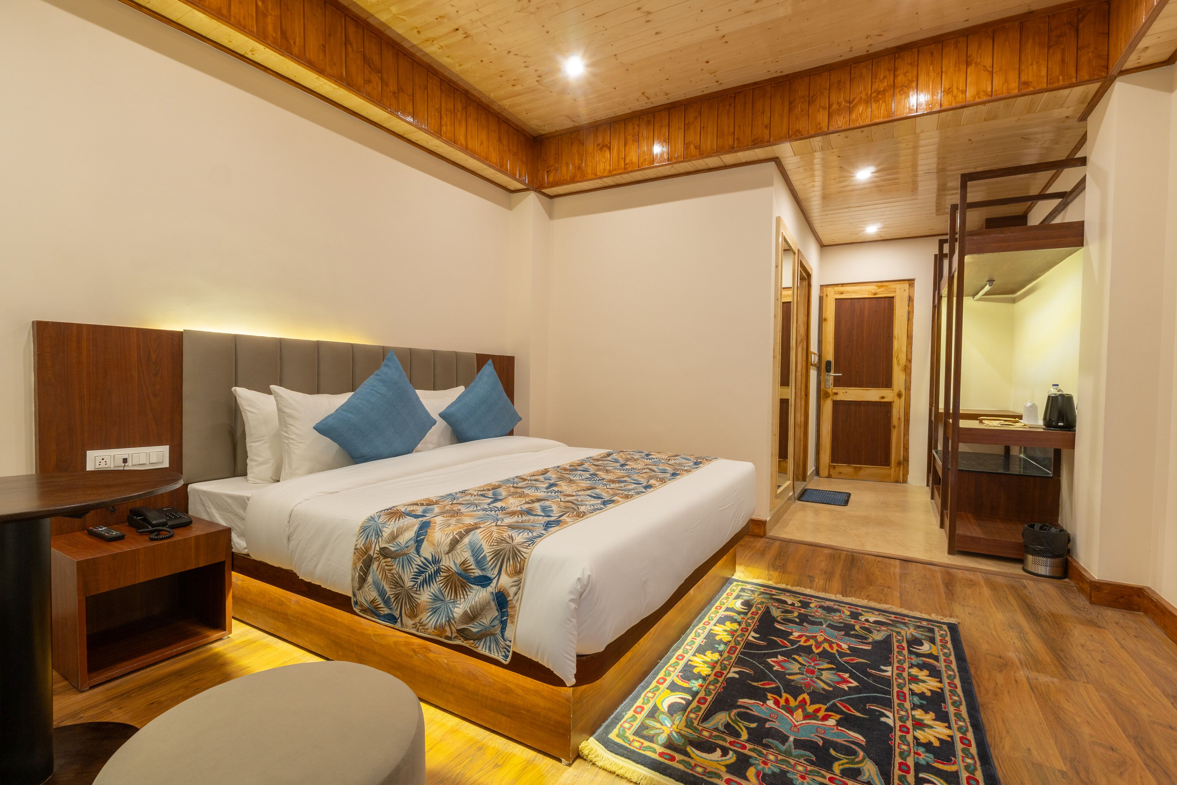 Une chambre moderne avec une tête de lit en bois, un grand lit avec une literie à motifs, une table de chevet avec un téléphone et un petit bureau.
