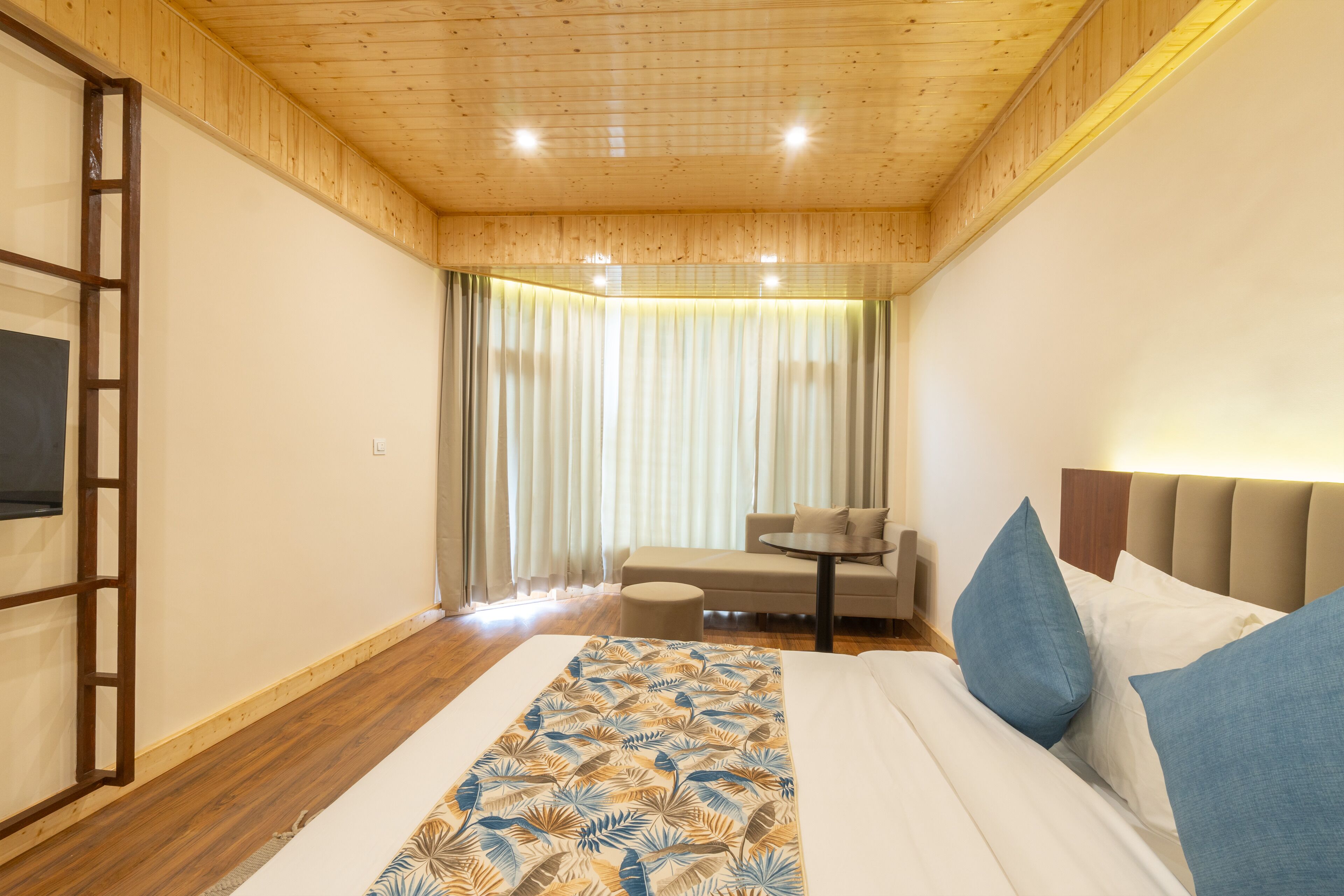 A hotel room with a wooden ceiling, a bed with a patterned bedspread, a small round table, a sofa, and a TV mounted on the wall.