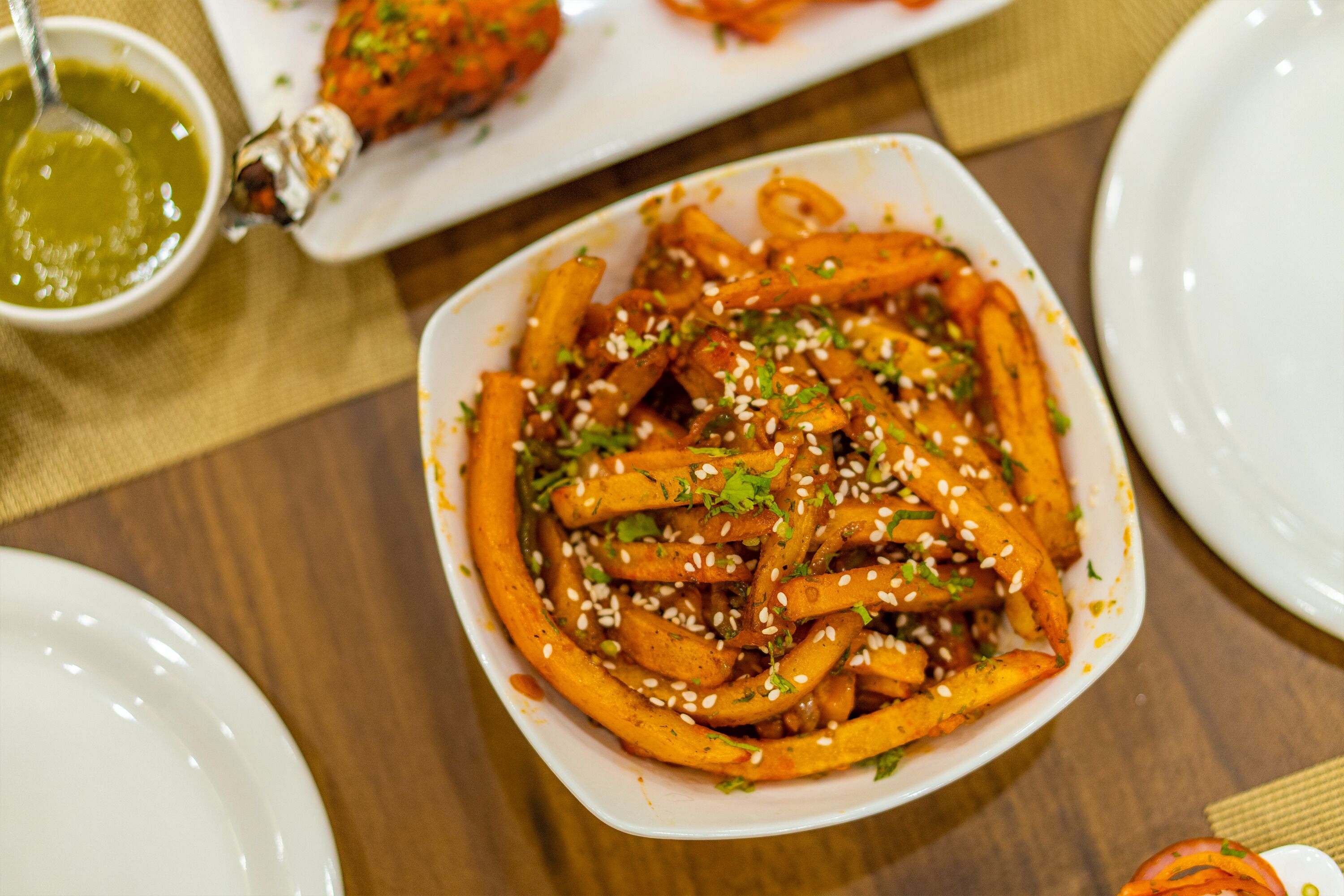 A bowl of seasoned fries garnished with sesame seeds and herbs, accompanied by a small bowl of green sauce.