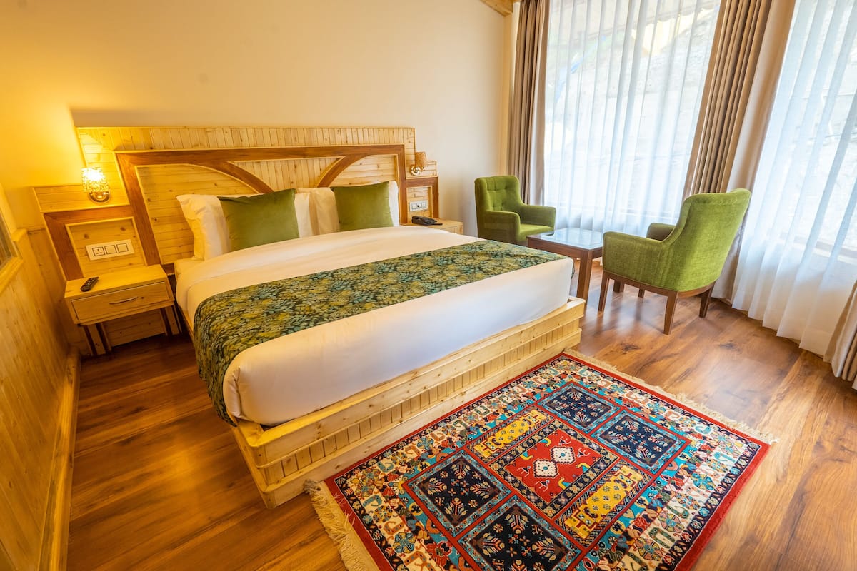 Een hotelkamer met een houten bed, groene stoelen, een kamerkleed met patroon en een nachtkastje met een lamp.