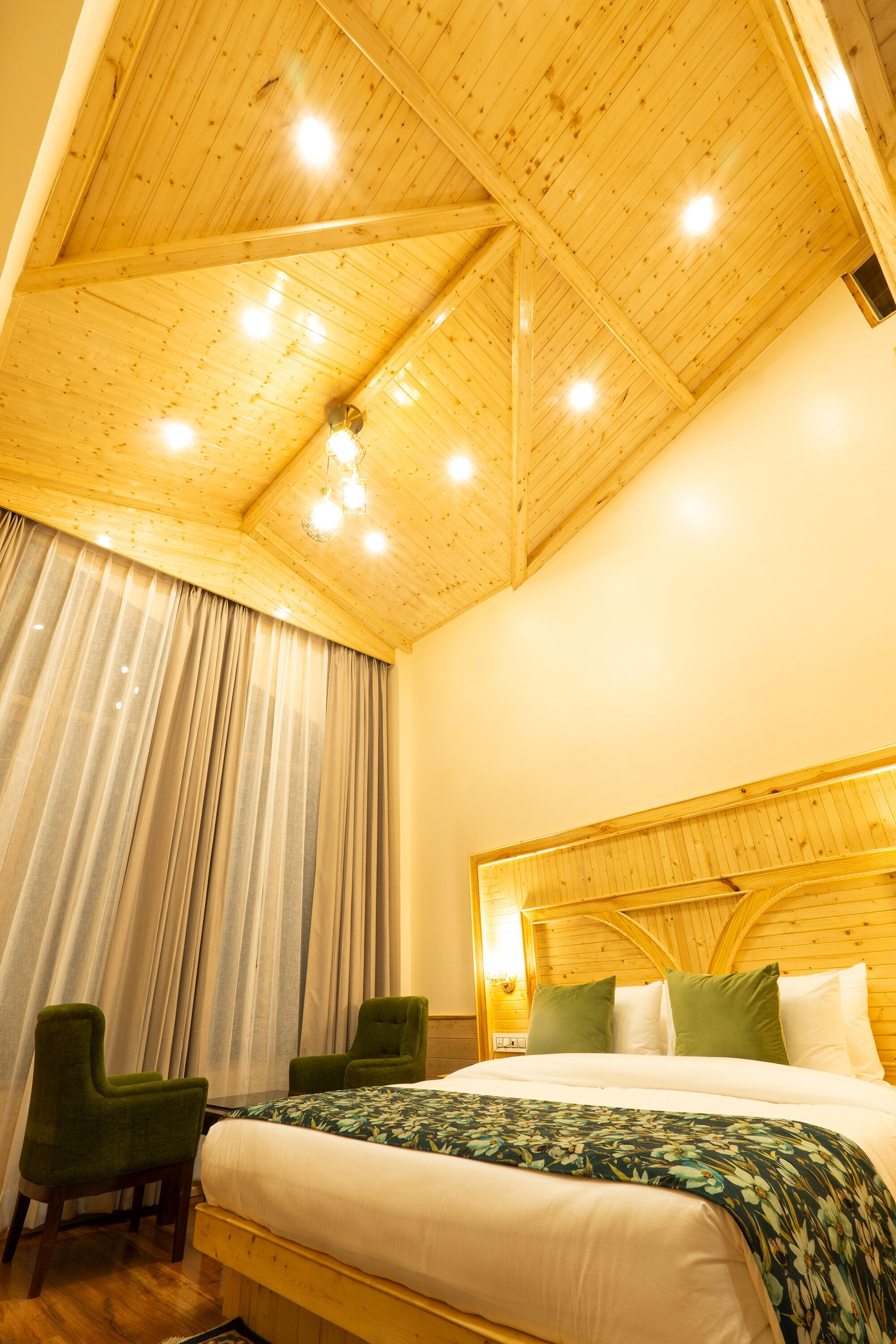 A bedroom with a wooden ceiling, a bed with green and white bedding, two green chairs, and a large window with curtains.