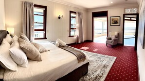 Un dormitorio amplio con una cama grande, dos ventanas con cortinas, una alfombra roja estampada, una silla y vistas al exterior.
