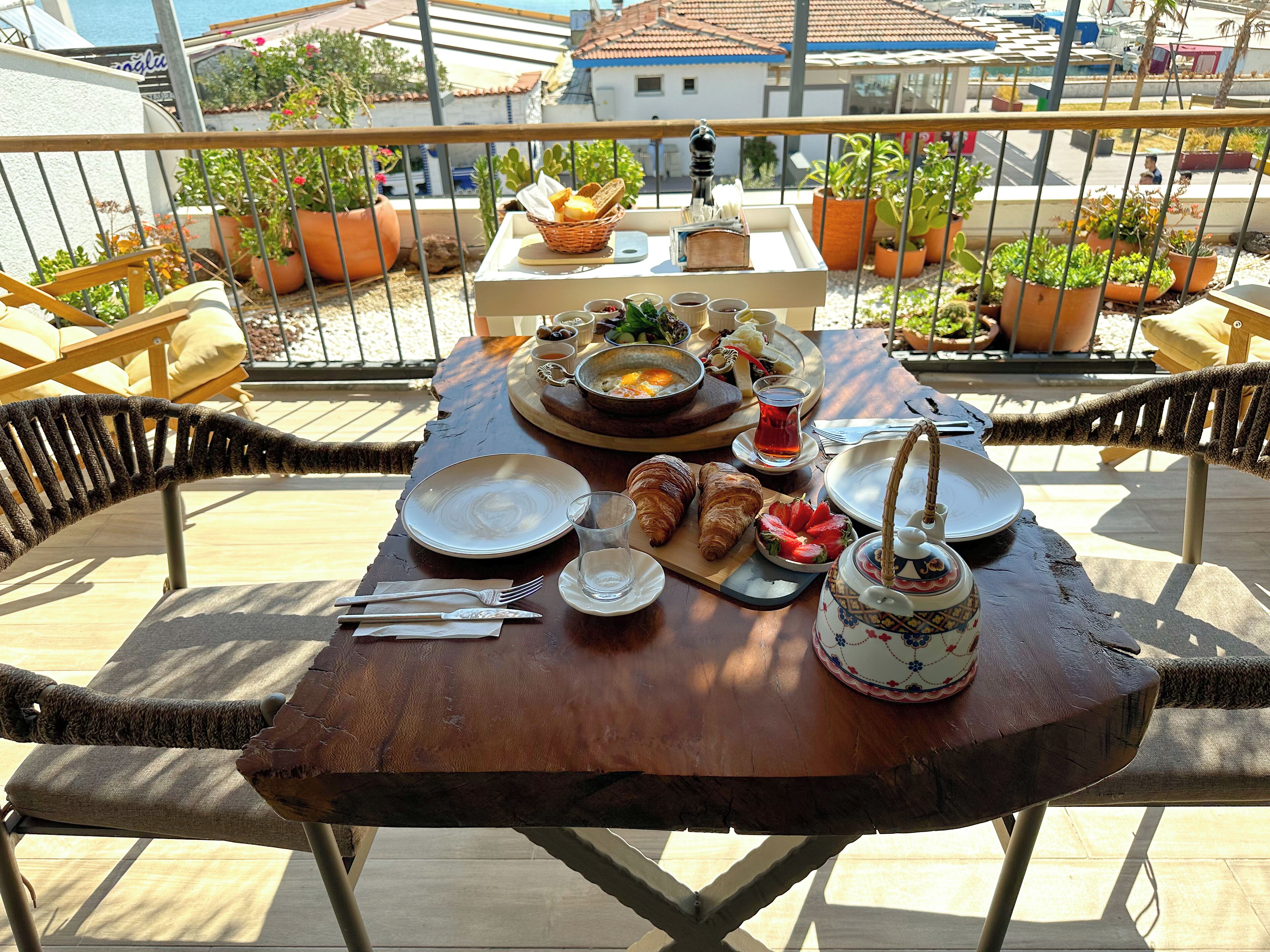 A wooden table set for breakfast with croissants, strawberries, and a teapot on a balcony overlooking a residential area.