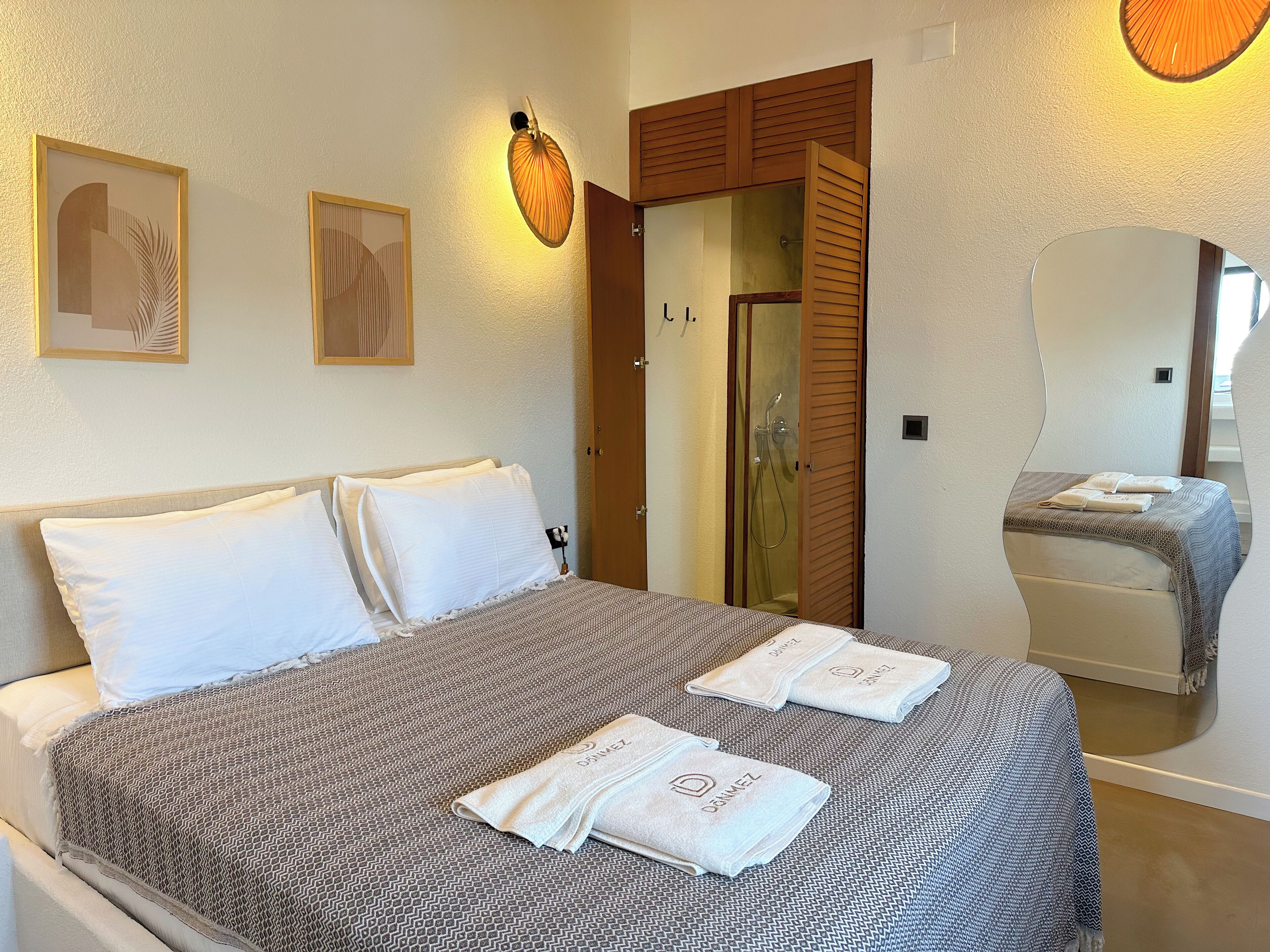 A neatly made bed with white pillows and a patterned blanket, a wooden door with a mirror, and two framed artworks on the wall.