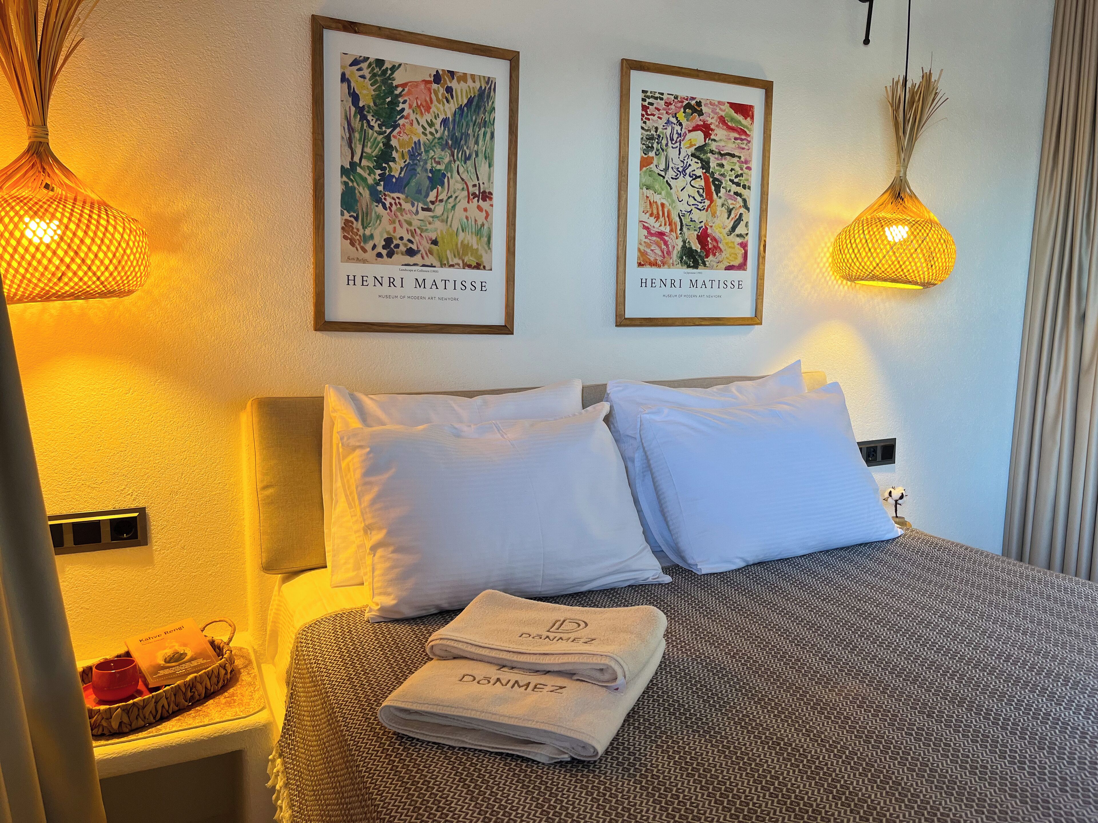 A hotel room with a bed, pillows, a framed artwork of Henri Matisse, and a small table with a candle and a card.