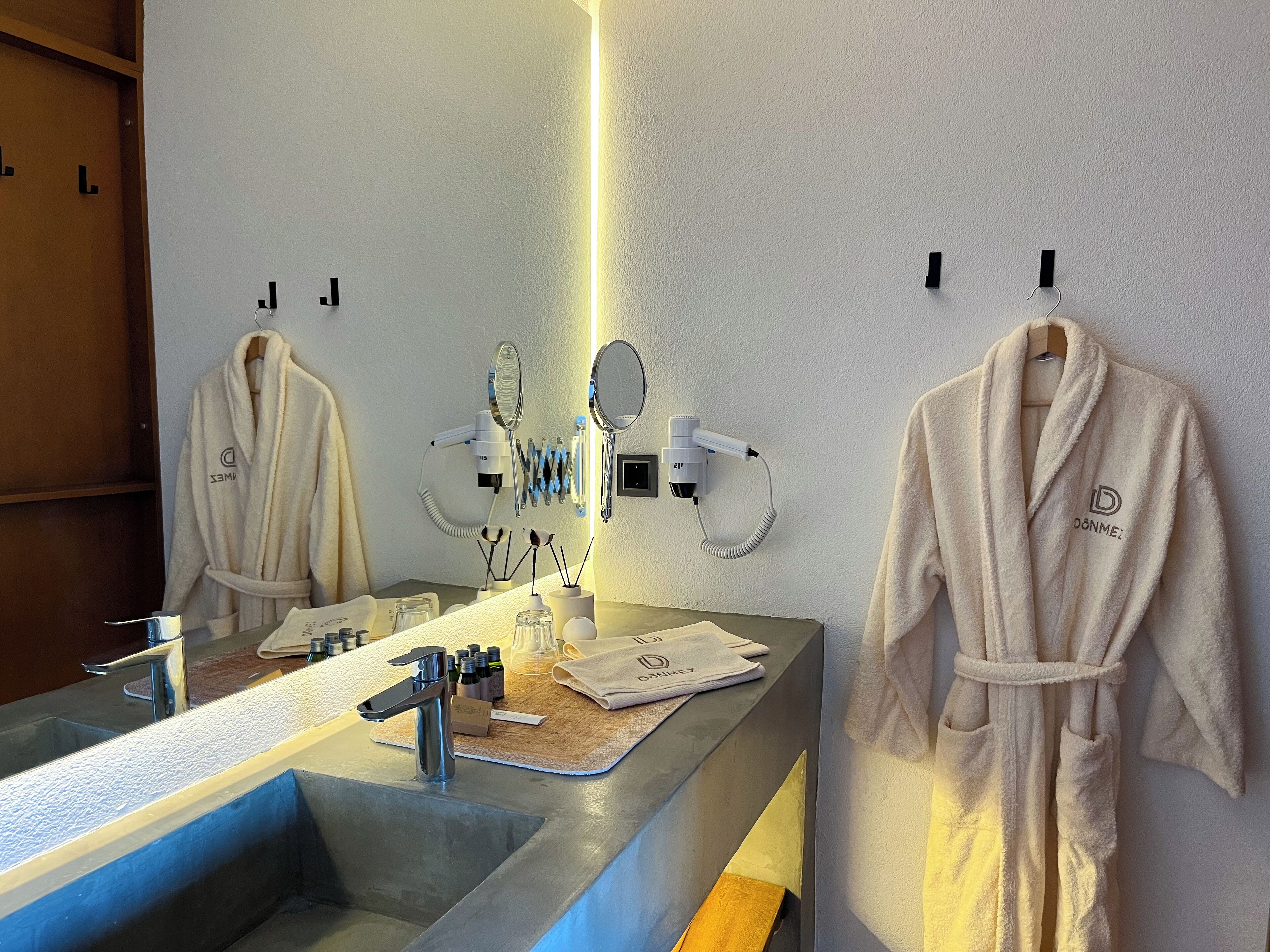 A hotel bathroom with a large mirror, two hair dryers, and two robes hanging on hooks.