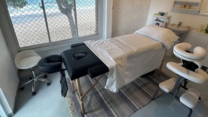 Een kamer met een bed, een massagetafel, een voetmassageapparaat, een klein krukje en een raam met uitzicht op het strand.