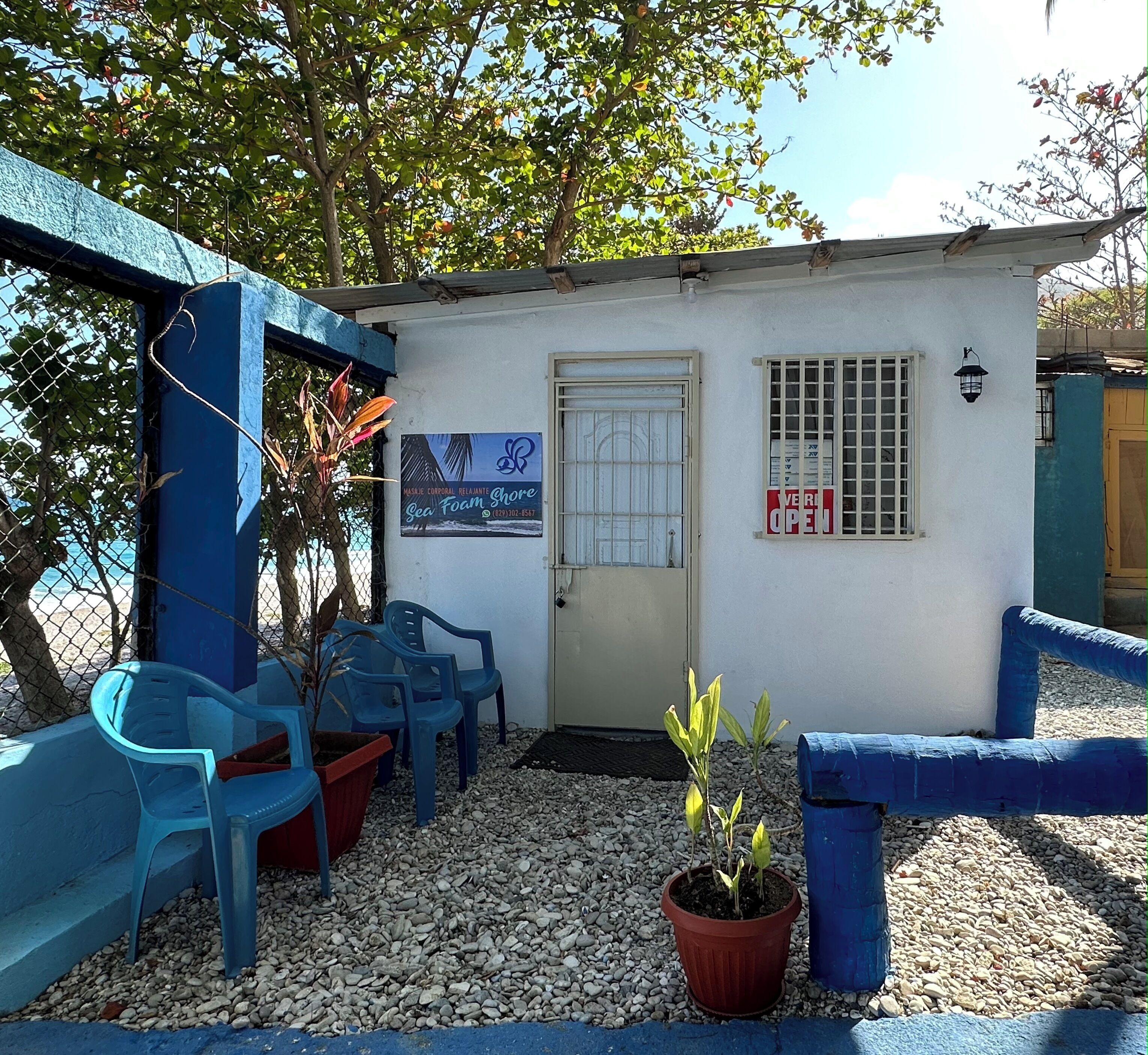 Un petit bâtiment avec une porte, une fenêtre grillagée et une enseigne indiquant qu’il est ouvert. On y voit des chaises bleues et un pot de fleurs devant l’entrée.