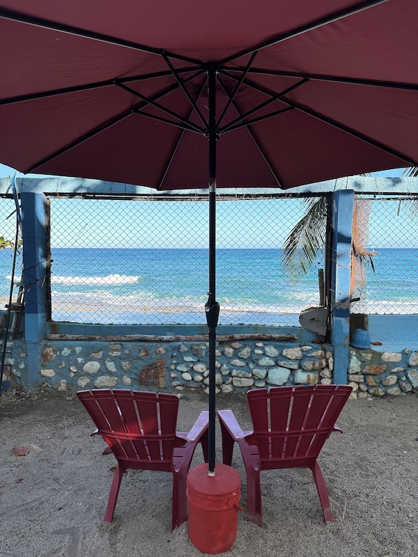 A view of the ocean from a covered patio with two red chairs and a large umbrella.