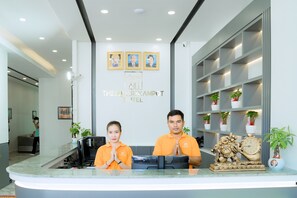 A reception desk with two staff members in orange uniforms, a reception area with a modern design, and a wall with framed pictures and the hotel name. - THE SQUARE KAMPOT HOTEL (Kampot)