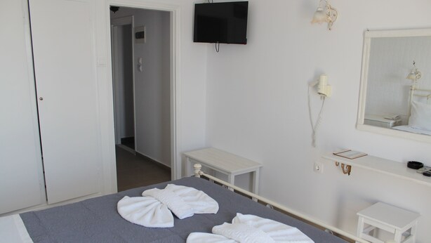 A hotel room with a bed, a television mounted on the wall, a bedside table, a mirror, and a small white cabinet. - Nikolas Hotel (Paros)
