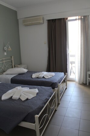 A hotel room with two beds, a bedside table, a lamp, and a window with curtains. - Nikolas Hotel (Paros)