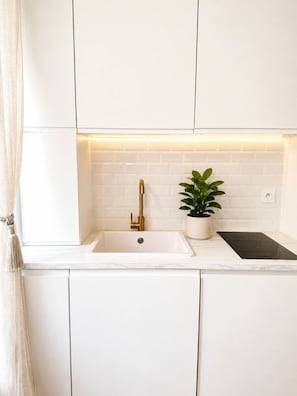 A kitchen with white cabinets, a gold faucet, a potted plant, and a black induction cooktop. - Cosy studio - Opera Garnier (Paris)