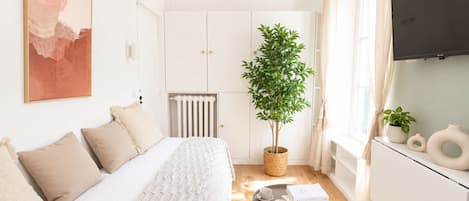 Un dormitorio moderno con cama, radiador, planta en maceta, una mesita con tazas y libros, y un televisor en la pared.