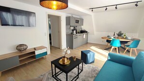 Una sala de estar moderna con un sofá, una mesa de centro y un televisor. Se observa una zona de cocina con una mesa de comedor y sillas.