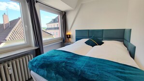 Un dormitorio con un ventanal grande, una cama con ropa de cama color turquesa y vistas de edificios.