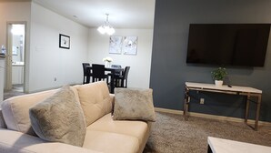 Ein Wohnzimmer mit beiger Couch, einem Esstisch mit Stühlen, einem an der Wand befestigten Flachbildfernseher und einem kleinen Tisch mit Pflanze.