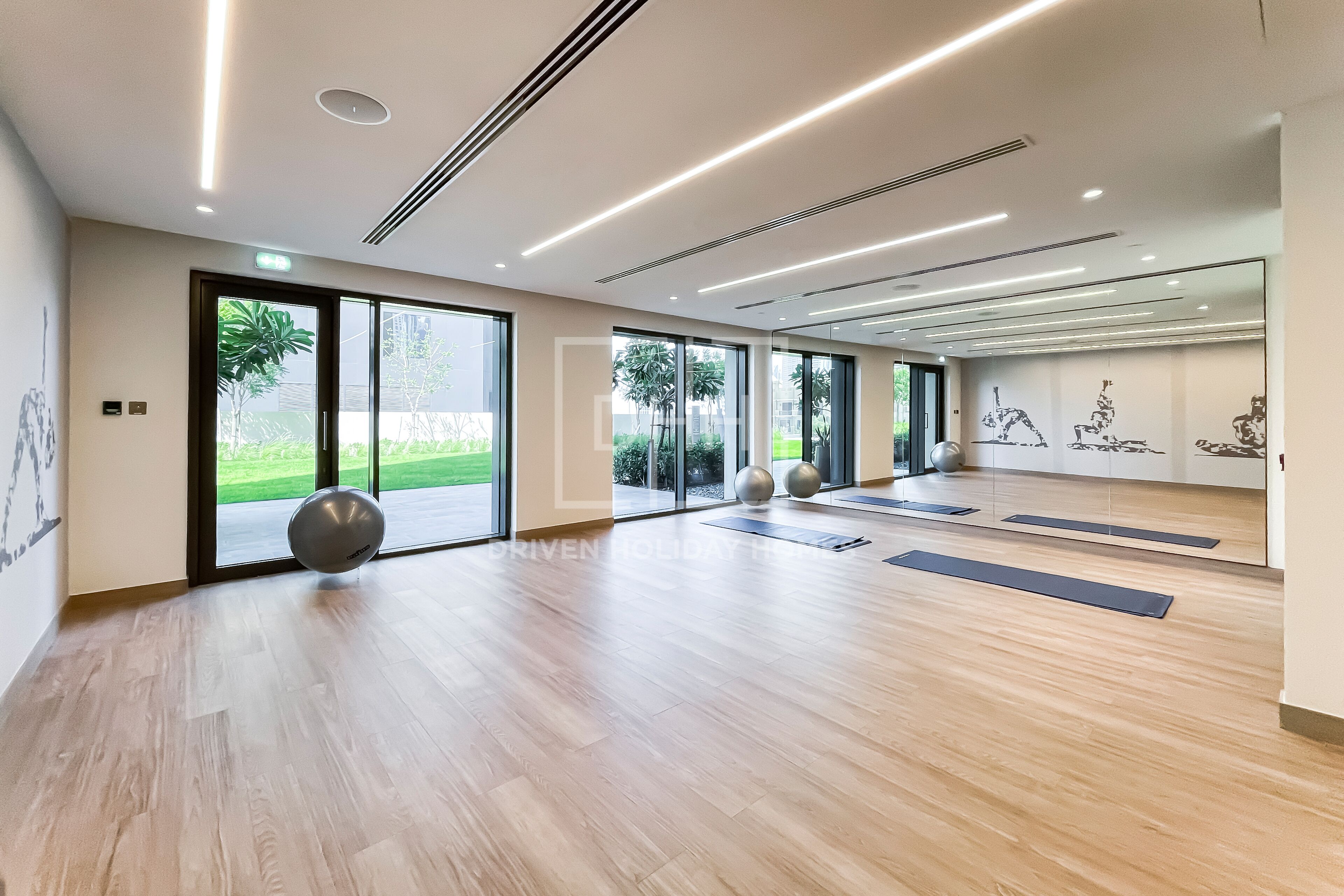 A spacious gym with wooden flooring, exercise mats, and large windows offering a view of greenery.