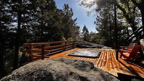 A wooden deck with a stone bench, surrounded by tall trees and a clear sky.
