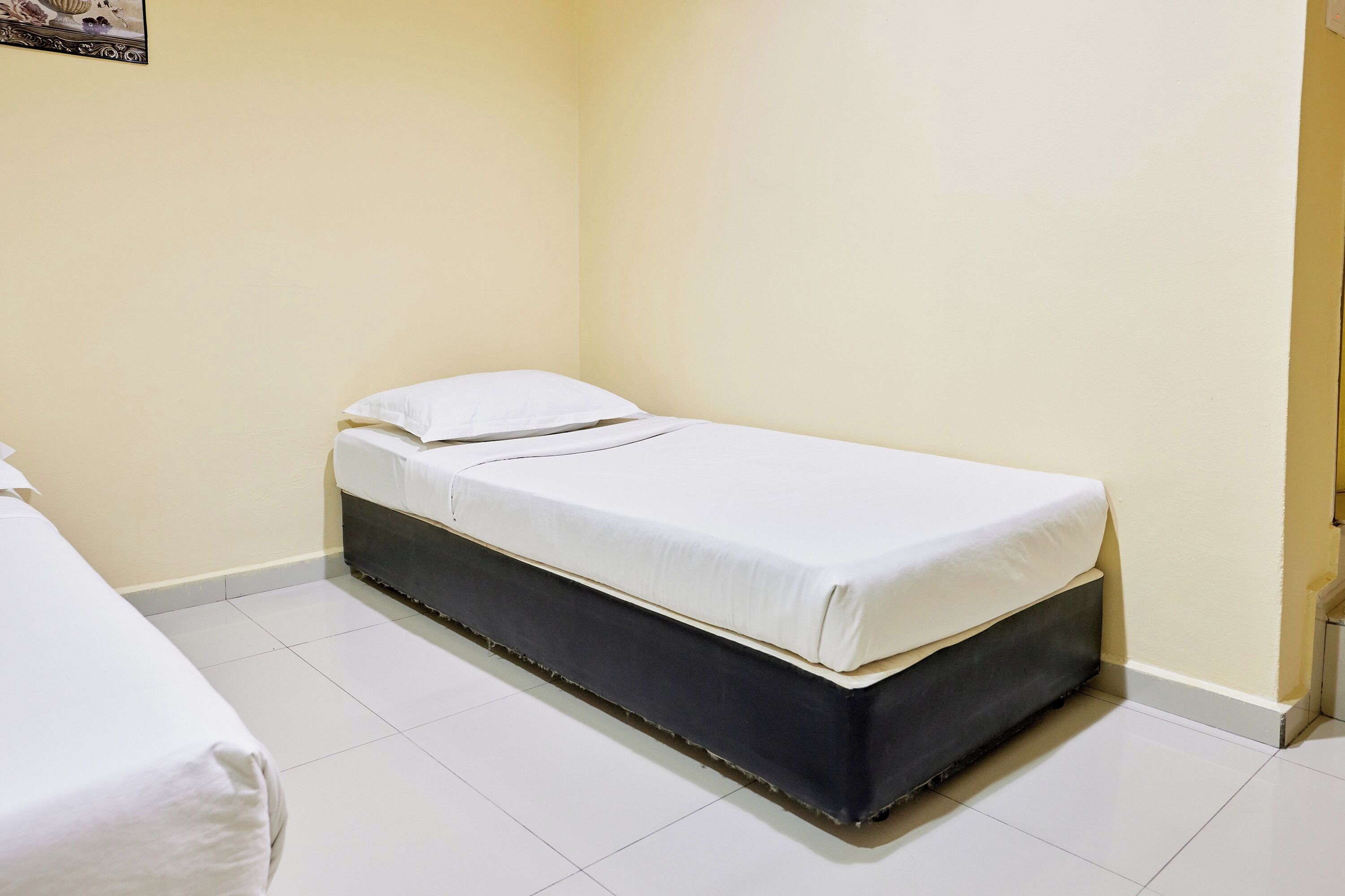 Ein kleines Zimmer mit einem Einzelbett, einer weißen Matratze und einem dunklen Bettgestell. Die Wände sind schlicht und der Boden ist mit Fliesen ausgelegt.