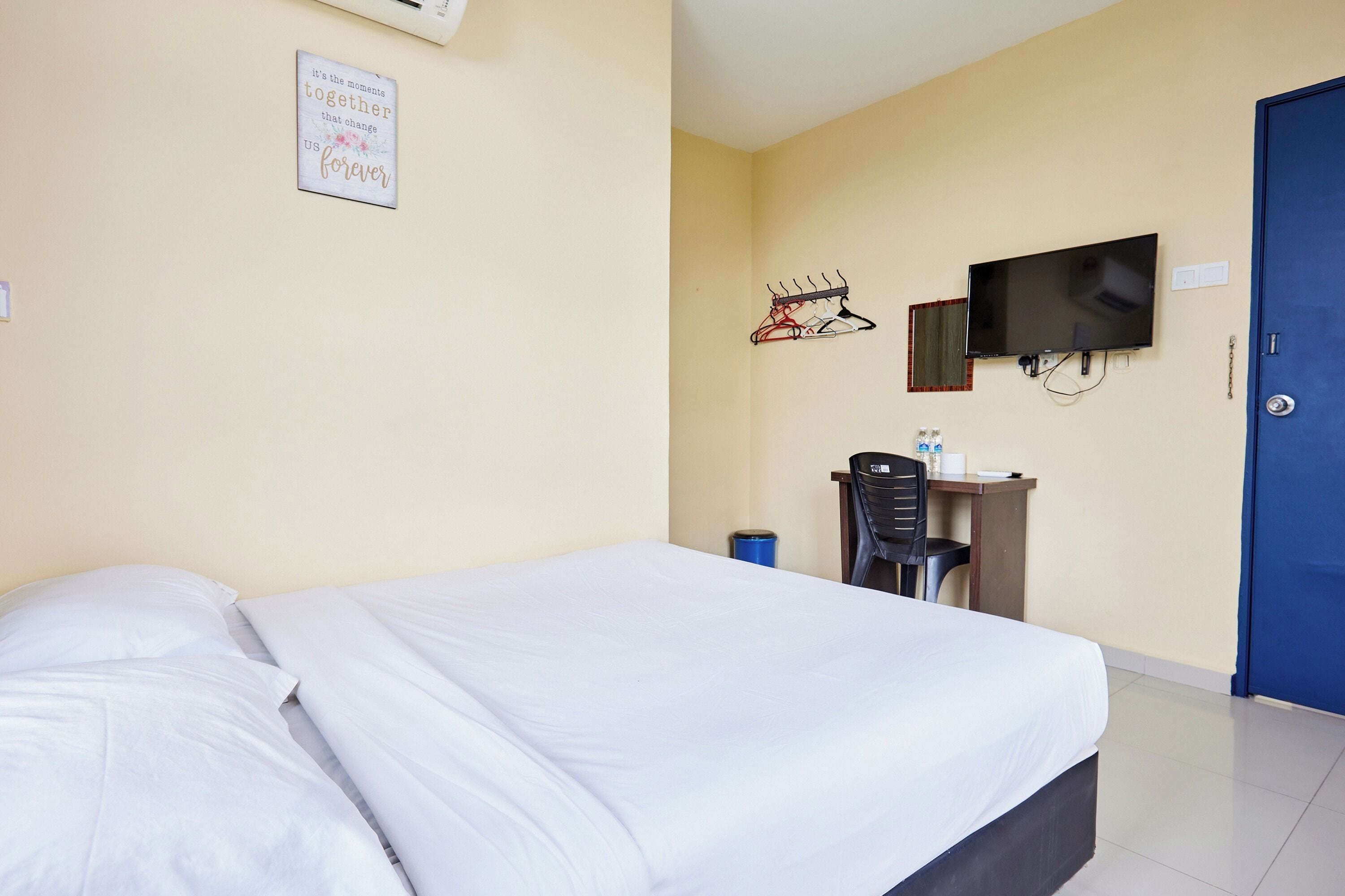 A hotel room with a bed, a desk with a chair, a TV, and a blue door.
