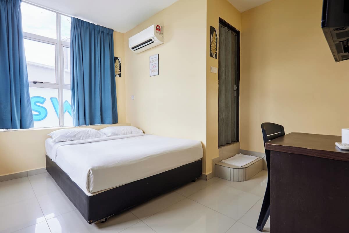 A hotel room with a bed, a desk, a chair, an air conditioner, a window with curtains, and a door.