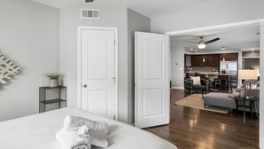 A modern bedroom with a neatly made bed, a small bedside table with a plant, and a white door leading to a kitchen area with a dining set.