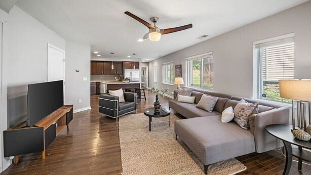 A spacious living room with a large sofa, a coffee table, and a TV. There is a kitchen in the background with modern appliances.